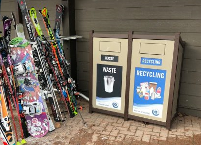 A new garbage bin beside a recycling bin. Both are located beside a ski rack covered in skis. 